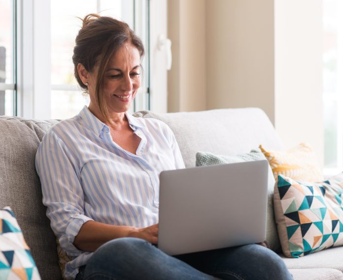 Middle aged woman using laptop in the sofa with a happy face standing and smiling with a confident smile showing teeth
